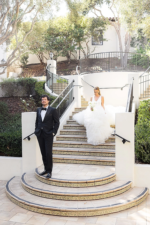 Wedding first look as bride descends tile stairs in a strapless ball gown, holding bouquet, toward groom in tux by white stucco courtyard