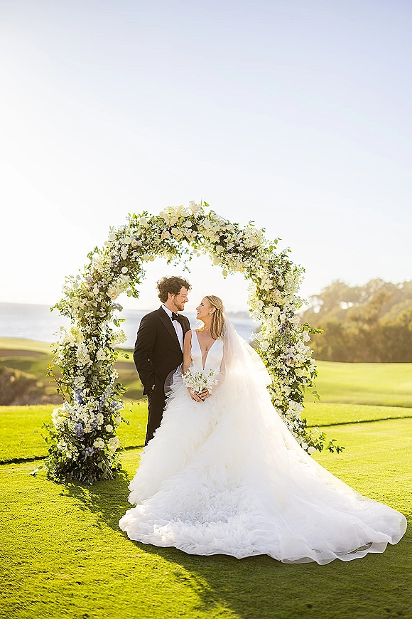 Couple portrait of bride in wedding dress and veil holding a bouquet beside groom in tuxedo under a floral arch with ocean view backdrop