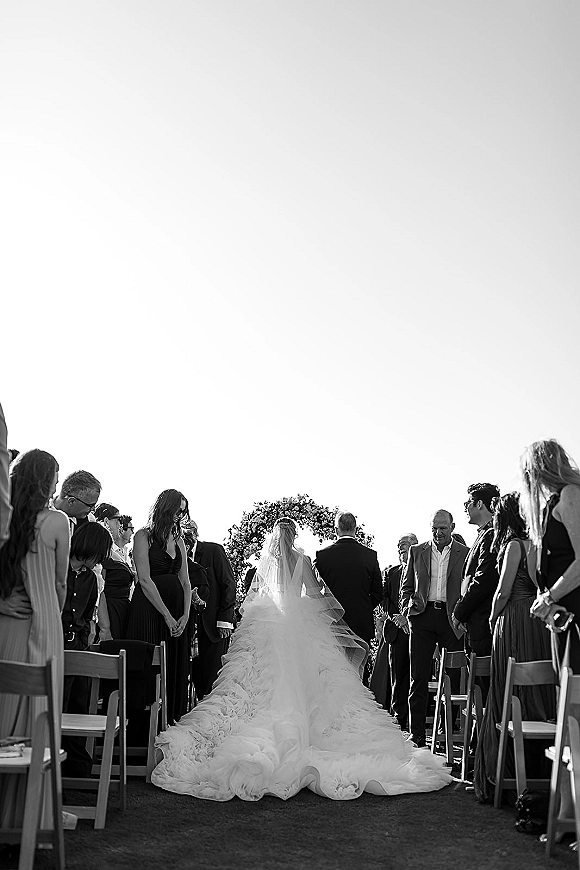 Wedding processional as bride walking down aisle, cathedral length veil and long train flowing toward a floral arch outdoors with guests standing