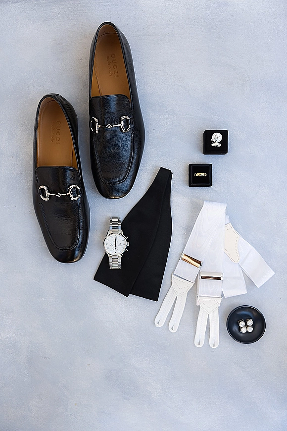 Groom accessories flat lay with black leather loafers, wristwatch, tie, cufflinks, and wedding band in a box on a light gray surface