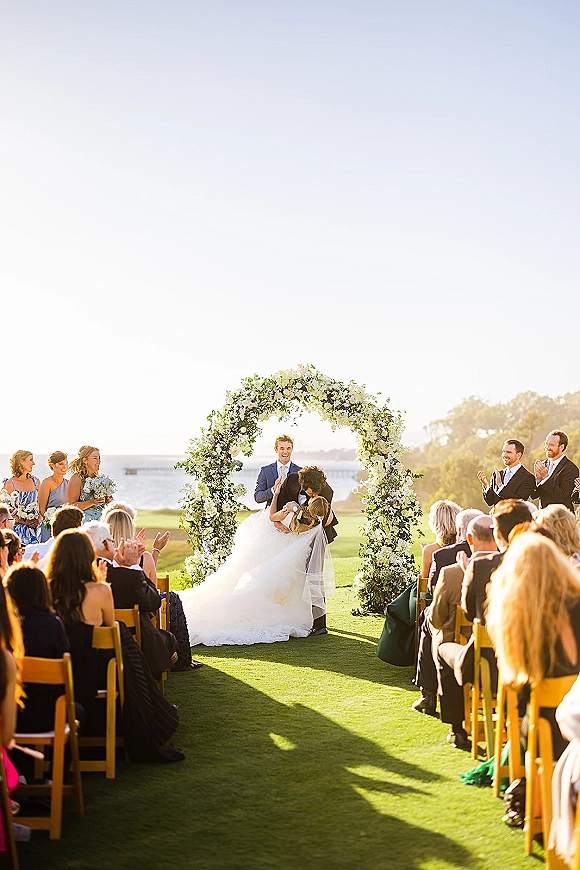 Ceremony kiss as bride in veil and groom in navy suit dip beneath white flower and greenery arch by the ocean, guests applauding