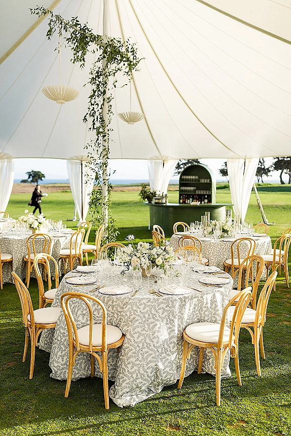Reception tablescape at an outdoor tented reception with patterned linens, white florals, bentwood chairs, and lanterns by the ocean horizon