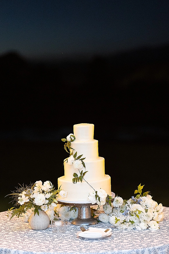 Wedding cake four tier wedding cake with smooth white buttercream, greenery garland and white roses on a silver stand against dark backdrop