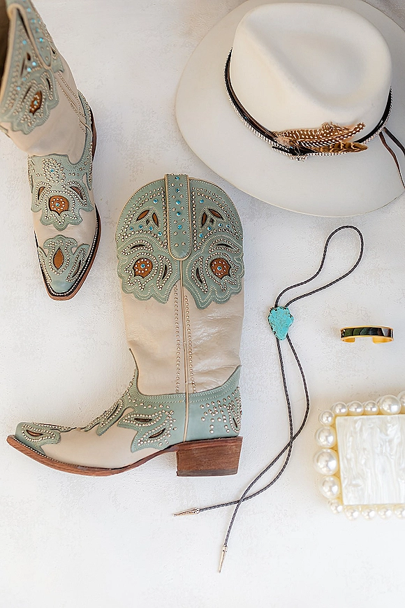 Groom accessories flatlay with western groom accessories, turquoise bolo tie, cowboy boots, white hat, gold wedding band on white surface