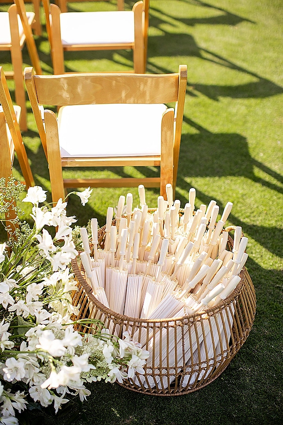 Ceremony fan basket with wedding ceremony fans, white paper hand fans beside white flowers and greenery on wooden chairs in sunlit lawn setting