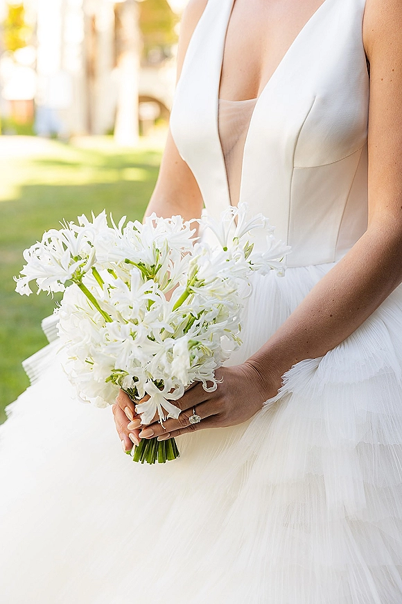 Bridal bouquet of white bridal bouquet blooms held against a deep V wedding gown, diamond engagement ring visible in sunlit lawn setting