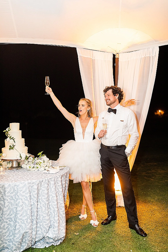 Wedding cake cutting as bride and groom toast with champagne flutes beside a tiered cake on a patterned table at night outdoors