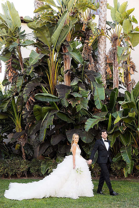 Couple portrait of bride and groom holding hands, her strapless tulle gown with long train and white bouquet amid palm greenery