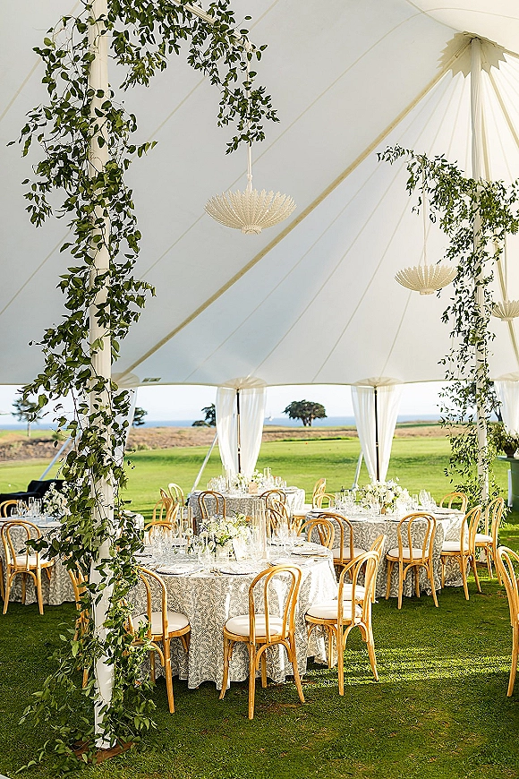Outdoor reception tent with sailcloth wedding tent chandeliers, greenery garlands, and round tables set on a lawn with ocean horizon beyond