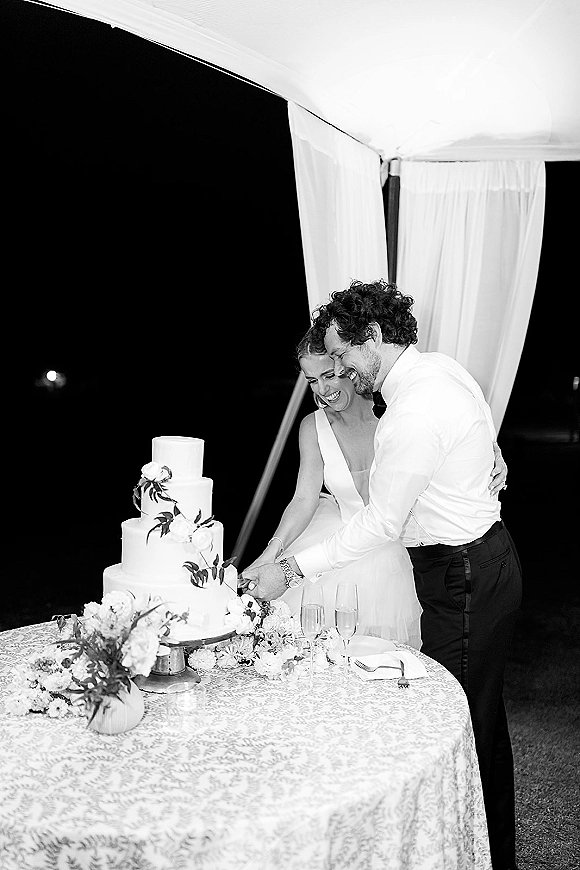 Wedding cake cutting as bride and groom hold a knife at a tiered cake with floral topper on a round table under tent draping at night