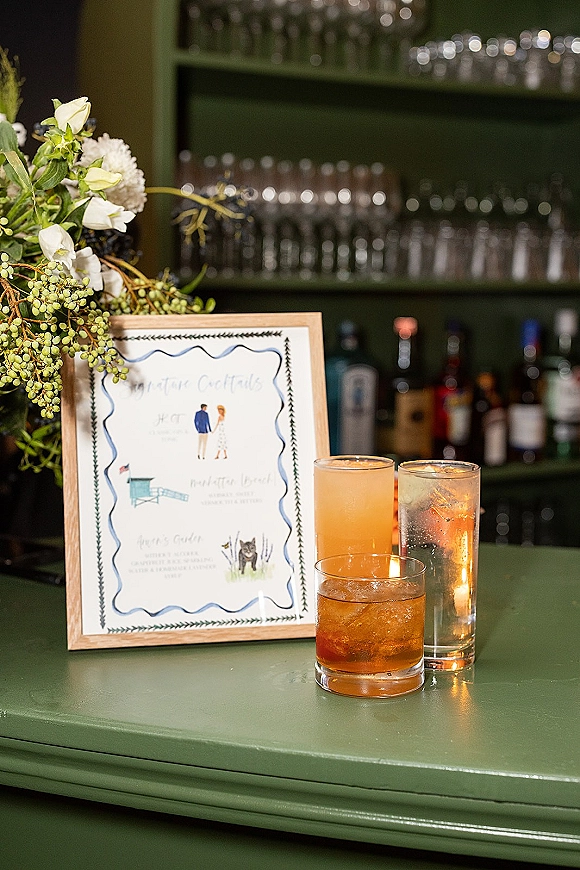 Signature cocktail sign in a framed display with floral greenery and a votive candle on a green bar cabinet with glasses and bottles