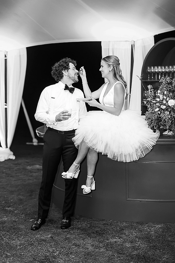 Cake cutting moment as bride in a tea length wedding dress feeds groom in bow tie and suspenders under white tent draping at night