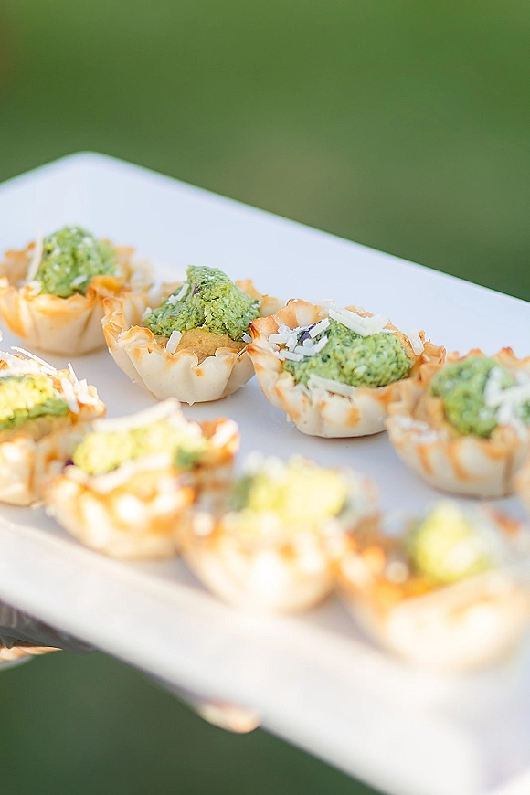 Wedding appetizers on a passed hors d'oeuvres tray, bite-size phyllo tartlets with green dip and shredded cheese garnish outdoors