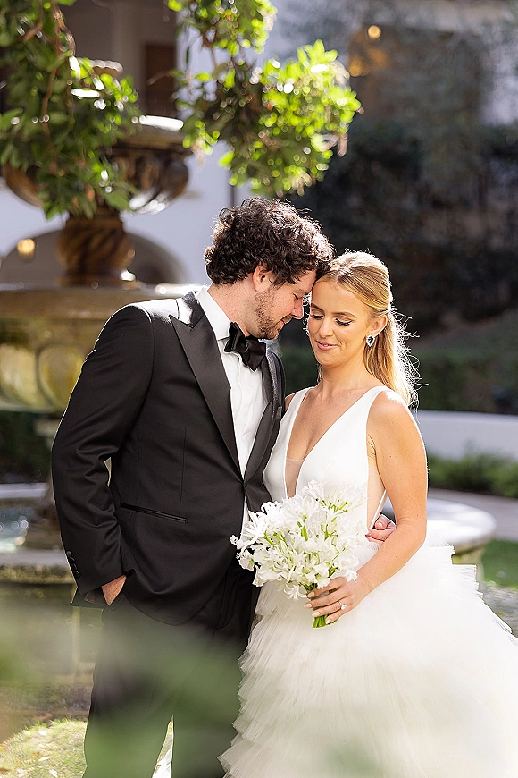 Couple portrait of bride and groom touching foreheads, bride holding white bouquet, groom in tuxedo by sunlit garden fountain