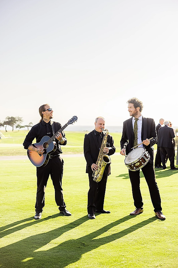 Wedding band performing live cocktail hour music with acoustic guitar accent on a grass lawn by the ocean horizon, guests watching nearby