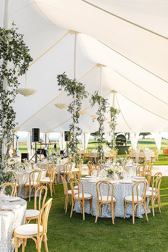Wedding reception tent with sailcloth tent wedding draping, round tables, greenery garlands, and hanging pendant lanterns on a lawn by water view