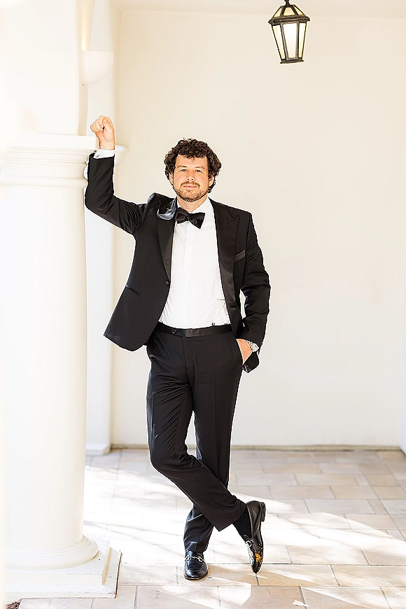 Groom portrait in a black tuxedo groom look, leaning on a white column under a covered walkway with lanterns and tiled floor