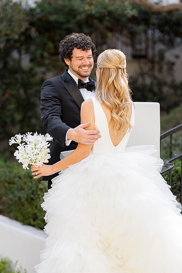Couple portrait of bride and groom embrace, groom holding bride in open-back tulle gown with white bouquet on garden steps