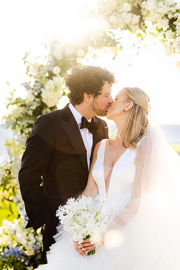 Wedding kiss portrait of bride and groom kiss under a floral arch, her veil trailing as she holds a white bouquet by the ocean sky backdrop