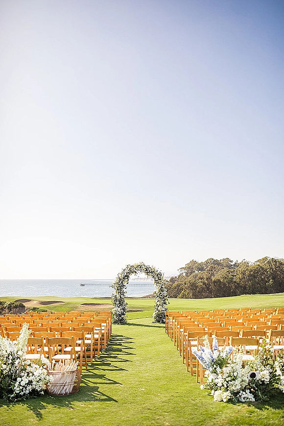 Outdoor ceremony setup with a wedding ceremony arch of white flowers and greenery, wooden chairs and aisle florals overlooking the ocean coastline