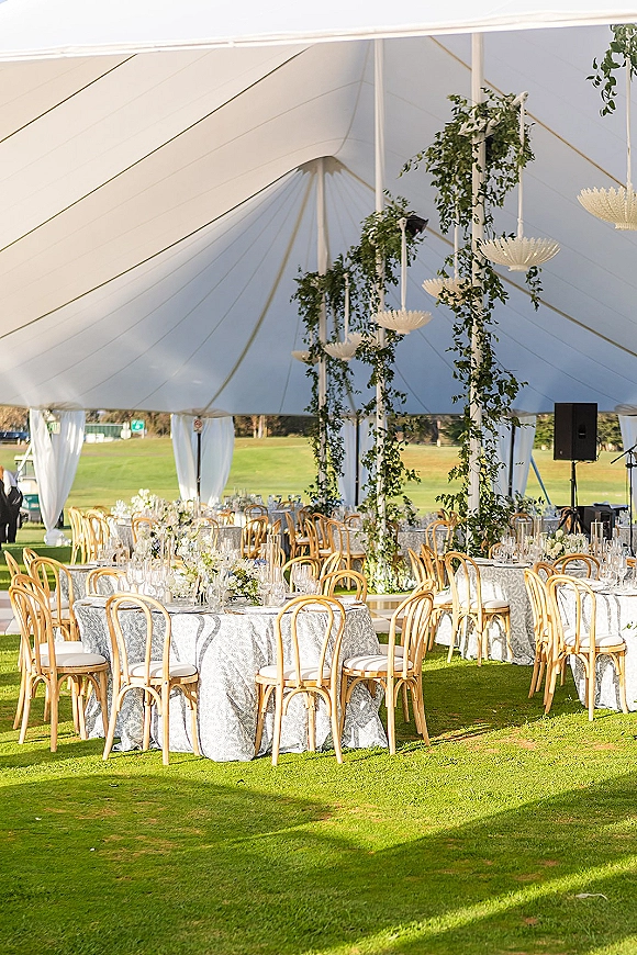 Reception tablescape with patterned linens, bentwood chairs, and white floral centerpieces beneath hanging greenery in a sailcloth tent on grass