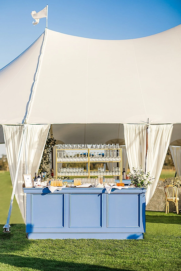 Wedding bar setup with a blue bar front, glassware and bottles on shelves, framed sign and florals under a white tent on lawn