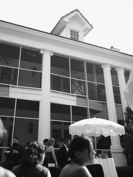 Wedding venue exterior with white column porch, patio umbrella and linen cocktail tables, and guests mingling beneath fans and lanterns