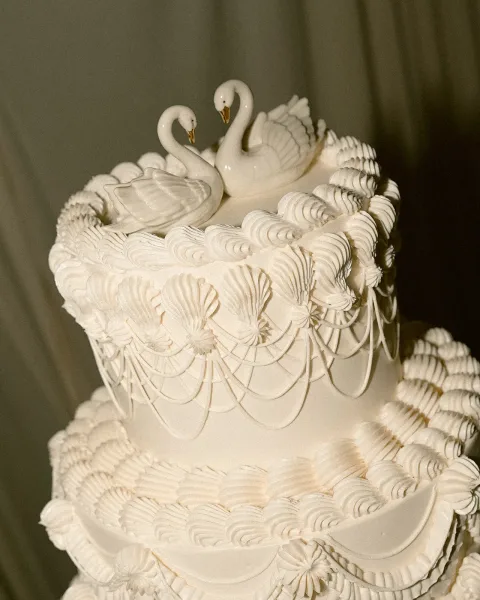 Wedding cake with white buttercream piped scallops and swan cake toppers, tiered with gold accents against a neutral backdrop