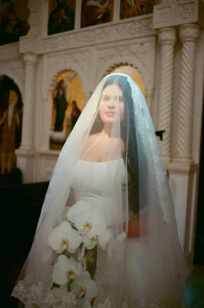 Bridal portrait of a bride with veil over her face, holding a white orchid bouquet in a church interior with religious icons and stone columns
