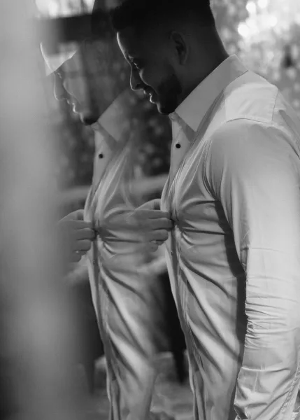 Groom getting ready, adjusting groom suspenders over a crisp dress shirt in a mirror-lit indoor room with soft window light