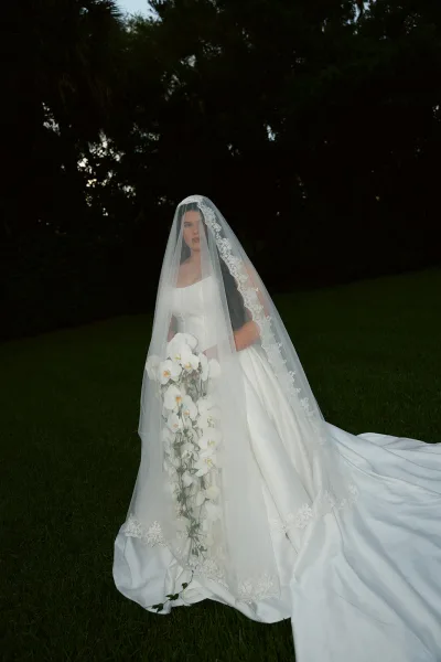 Bridal portrait of a cathedral veil bride holding a cascading white orchid bouquet, strapless gown with long train on a lawn at dusk