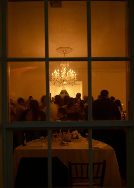 Wedding reception with a crystal chandelier over a round table set with white tablecloth, floral centerpiece, candles, and guests beyond window panes