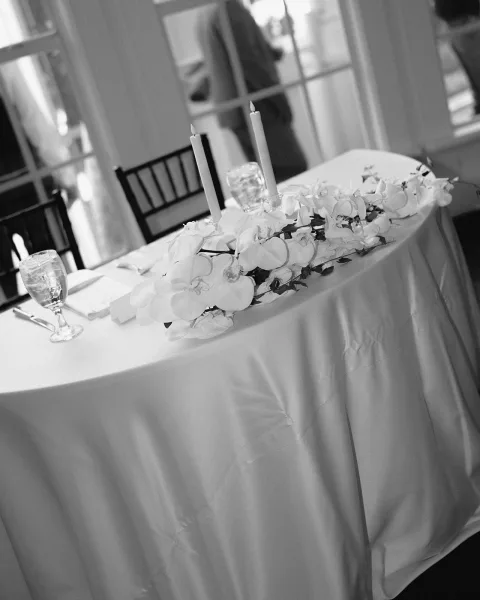 Sweetheart table decor with an orchid table runner, taper candles in glass holders, white linens, and black chiavari chair by glass doors