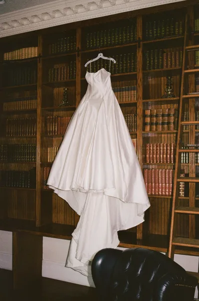 Wedding dress, a strapless bridal gown on a hanger in a wood-paneled library with built-in bookshelves, ladder, and leather chair