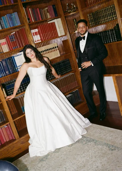Couple portrait of bride in a strapless satin ball gown with long train and groom in black tuxedo holding a cocktail, posed by library bookshelves