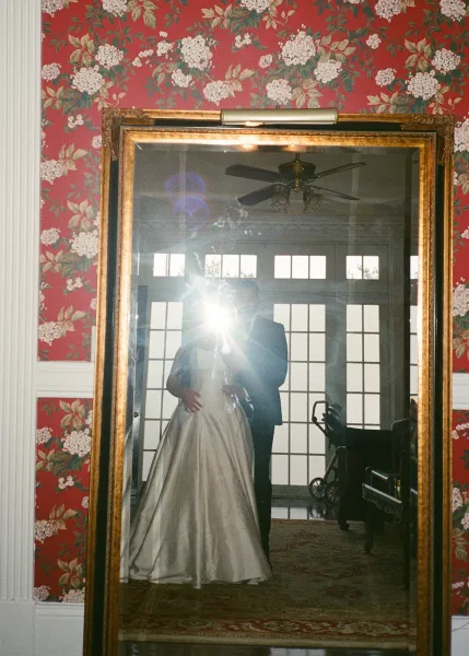 Couple portrait in a gold framed mirror, bride in a strapless satin wedding dress hugging groom in tuxedo against floral wallpaper