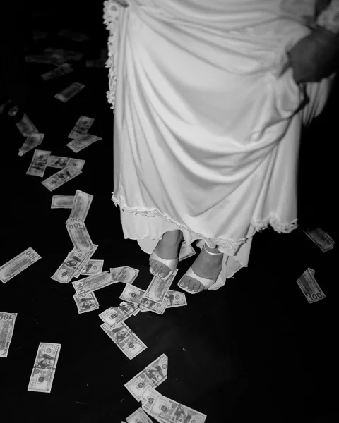 Bridal shoe detail with white bridal sandals peeking from the wedding dress hem, scattered cash around her feet on a dark floor