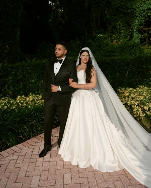 Couple portrait of bride holding groom’s arm in a strapless gown and lace veil train beside a brick walkway with garden greenery