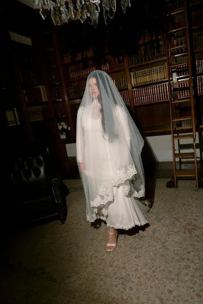 Bridal portrait of a bride in veil wearing a long-sleeve lace dress and ankle-strap heels in a library with bookshelves and ladder