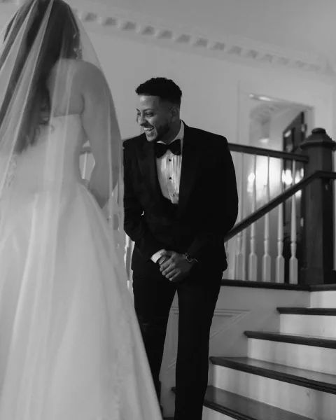 Wedding first look as bride in a strapless ball gown and long veil approaches laughing groom in black tuxedo on an indoor staircase