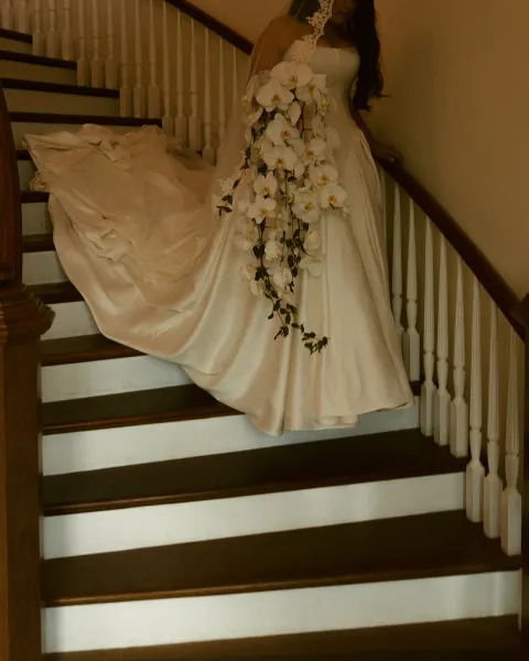 Bridal portrait of a bride on staircase in a strapless wedding dress train, lace veil, holding a cascading white orchid bouquet by a wood stair railing