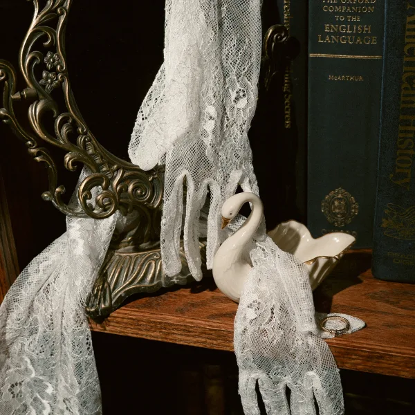 Wedding ring flatlay with engagement ring close-up resting on lace bridal gloves beside an ornate mirror frame, books, and ceramic swan on dark shelf