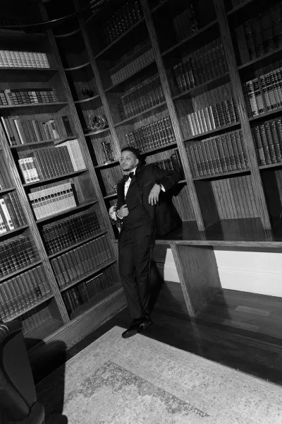 Groom portrait in a tuxedo with bow tie and boutonniere, holding a bouquet beside library bookshelves and wood paneling indoors