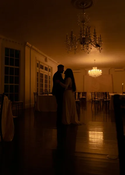 First dance of bride in a wedding dress and groom in a suit beneath crystal chandeliers, candlelit ballroom with tall windows and wood floor