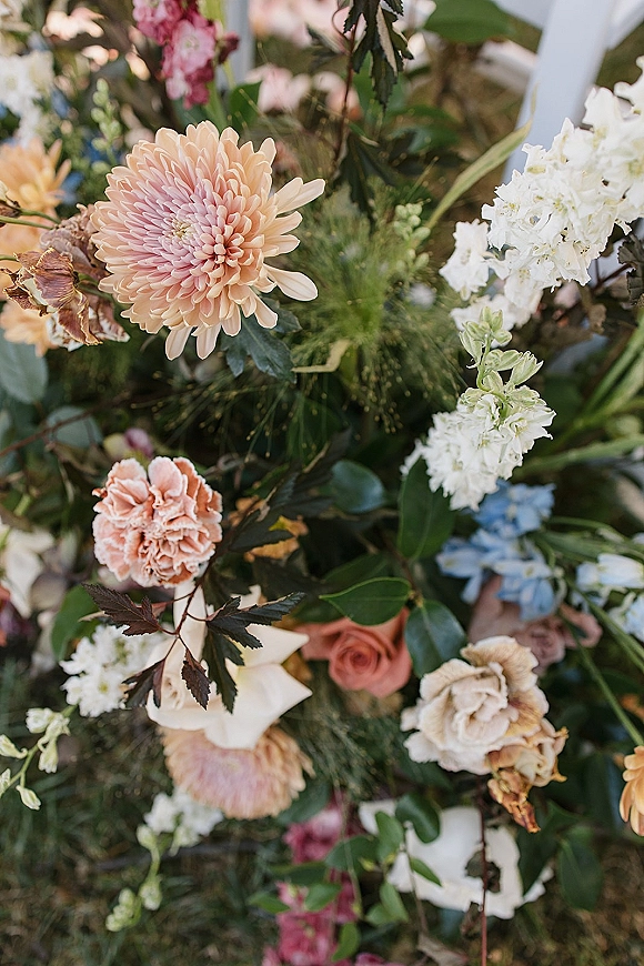 Wedding floral arrangement of peach and blush dahlias, roses, hydrangea and greenery draped on a white chair in a garden setting