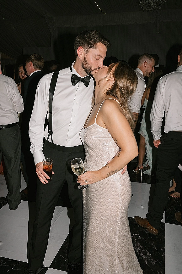 Wedding kiss on the reception dance floor as bride in a sparkly dress and groom in suspenders hold drinks under string lights