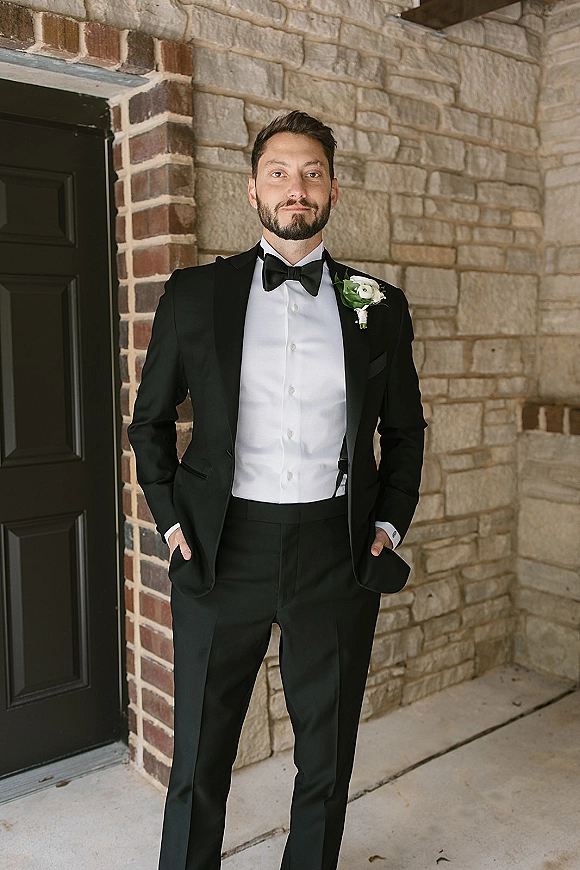 Groom portrait in a black tuxedo, bow tie, and white rose boutonniere, hands in pockets by a stone wall and brick doorway