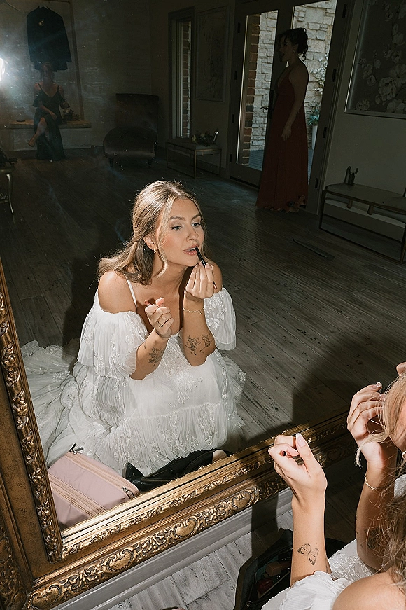 Bridal makeup as bride applying lipstick in an ornate mirror, wearing an off-shoulder wedding dress with earrings in a getting ready room