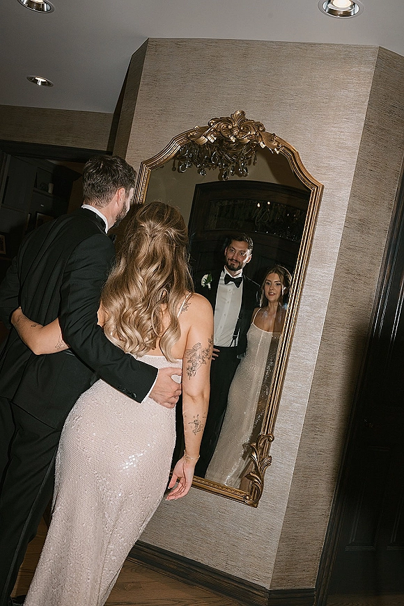 Couple portrait reflected in an ornate gold mirror, bride in sequin dress and groom in tuxedo under a chandelier in a hallway