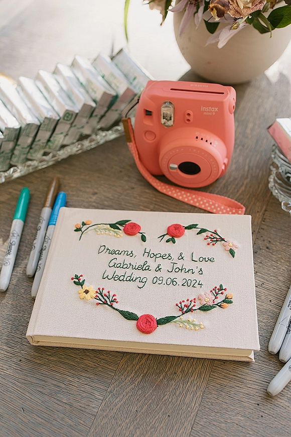 Wedding guest book with floral embroidery on a wood table beside a coral instant camera, film packs, markers, and flowers